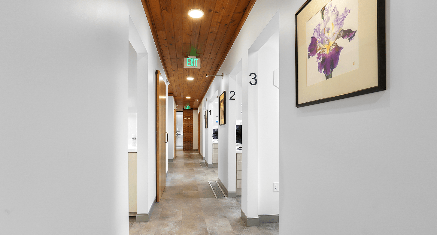 A clean, modern hallway with numbered doors and a painting of purple irises. Wood ceiling and white walls create a calm, professional atmosphere.