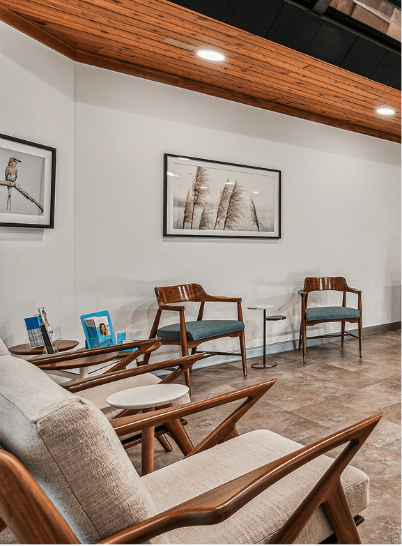 Modern waiting area with wooden chairs and teal cushions, small round tables, and nature artwork on white walls. Warm lighting and calm ambiance.