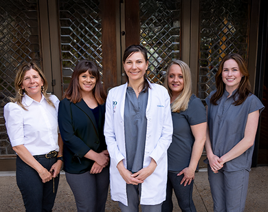 A group of five women, four in gray attire and one in a white coat, smile warmly while standing in front of a decorative glass-paneled door.