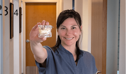 A smiling person in scrubs holds a dental mold in a hallway, conveying professionalism and friendliness in a clinical setting.