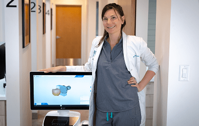 Dr. Patt in a lab coat stands beside a computer displaying a dental 3D model in a modern clinic hallway.