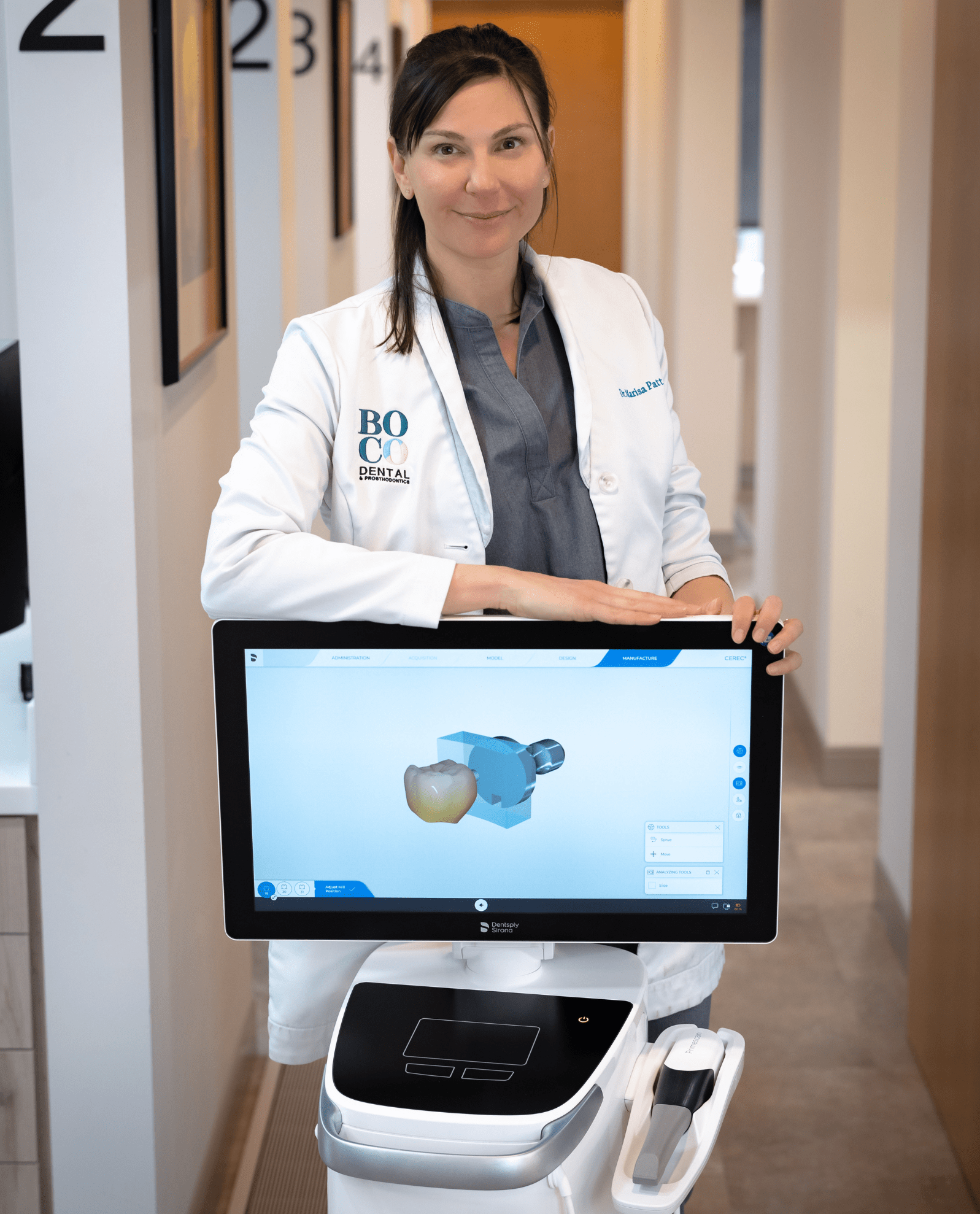 A dental professional stands beside a modern digital device displaying a 3D model of a tooth. The clinic has a modern and clean interior.