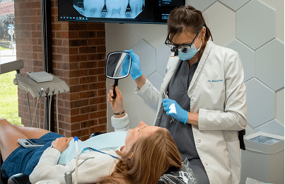 Dr.Patt shows a patient their reflection in a mirror while discussing dental care, surrounded by modern equipment in a bright office.