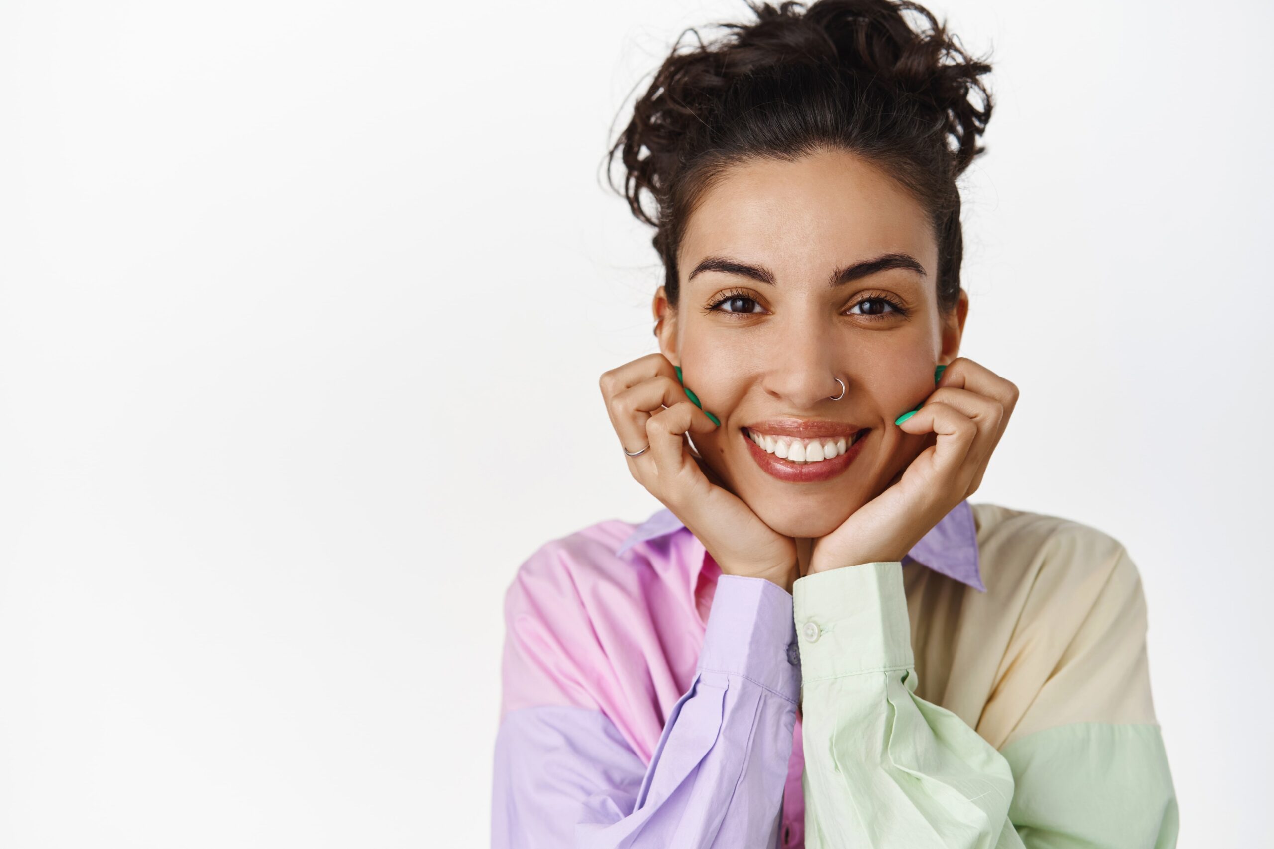 Smiling woman with curly hair holds her chin with both hands. She wears a colorful pastel shirt and has a nose ring, conveying happiness and warmth.
