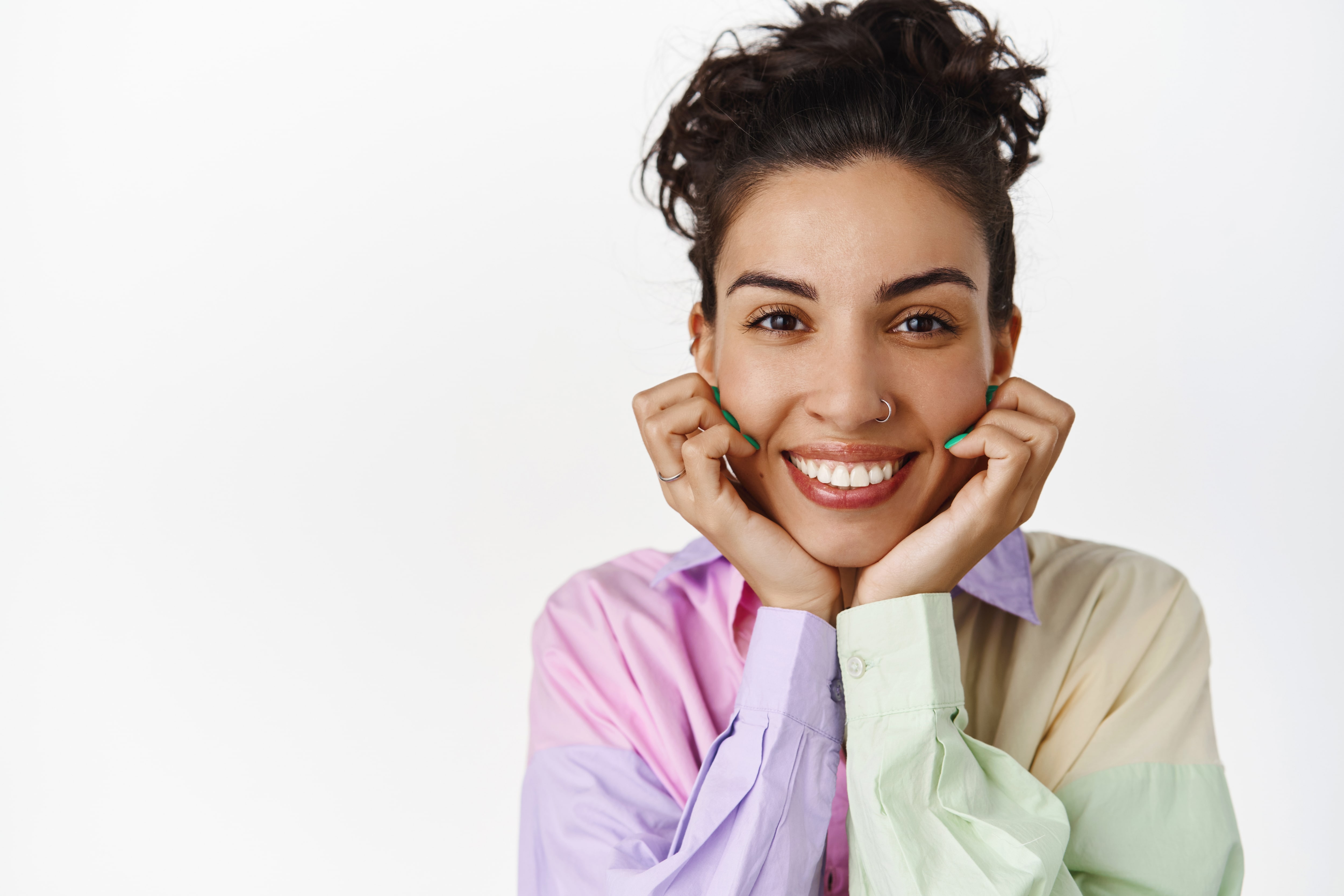 Smiling woman with curly hair holds her chin with both hands. She wears a colorful pastel shirt and has a nose ring, conveying happiness and warmth.