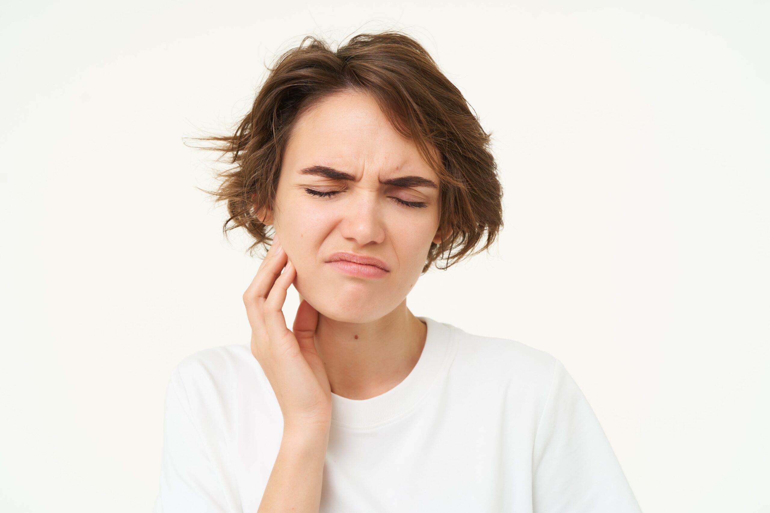 A person with short brown hair in a white shirt touches their cheek with a pained expression, indicating discomfort or toothache. White background.