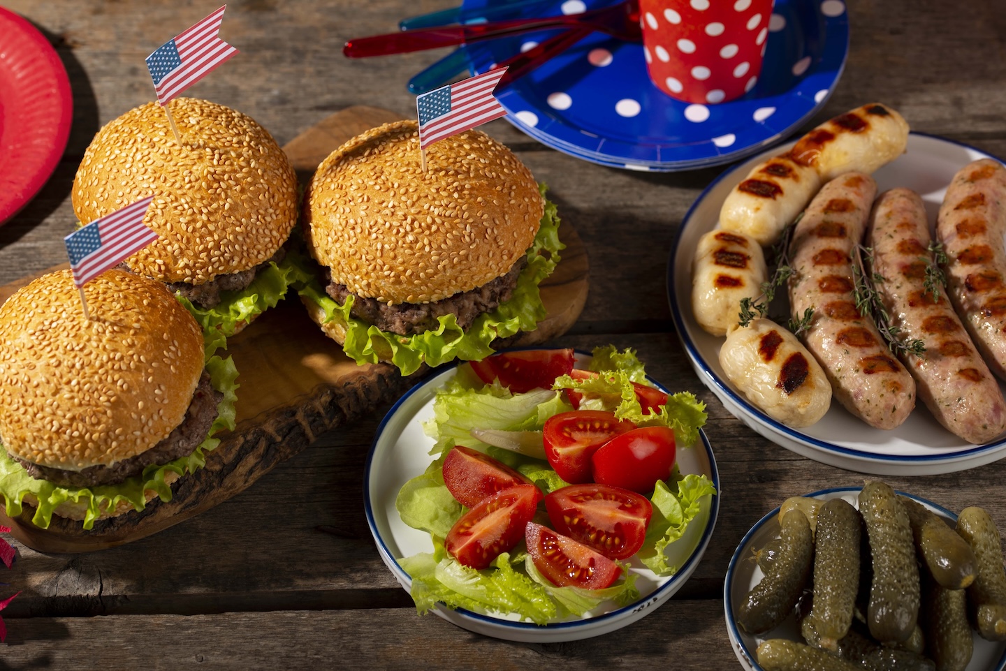 A picnic table with three burgers topped with American flags, grilled sausages, a salad of lettuce and tomatoes, and a side of pickles. Festive and summery.