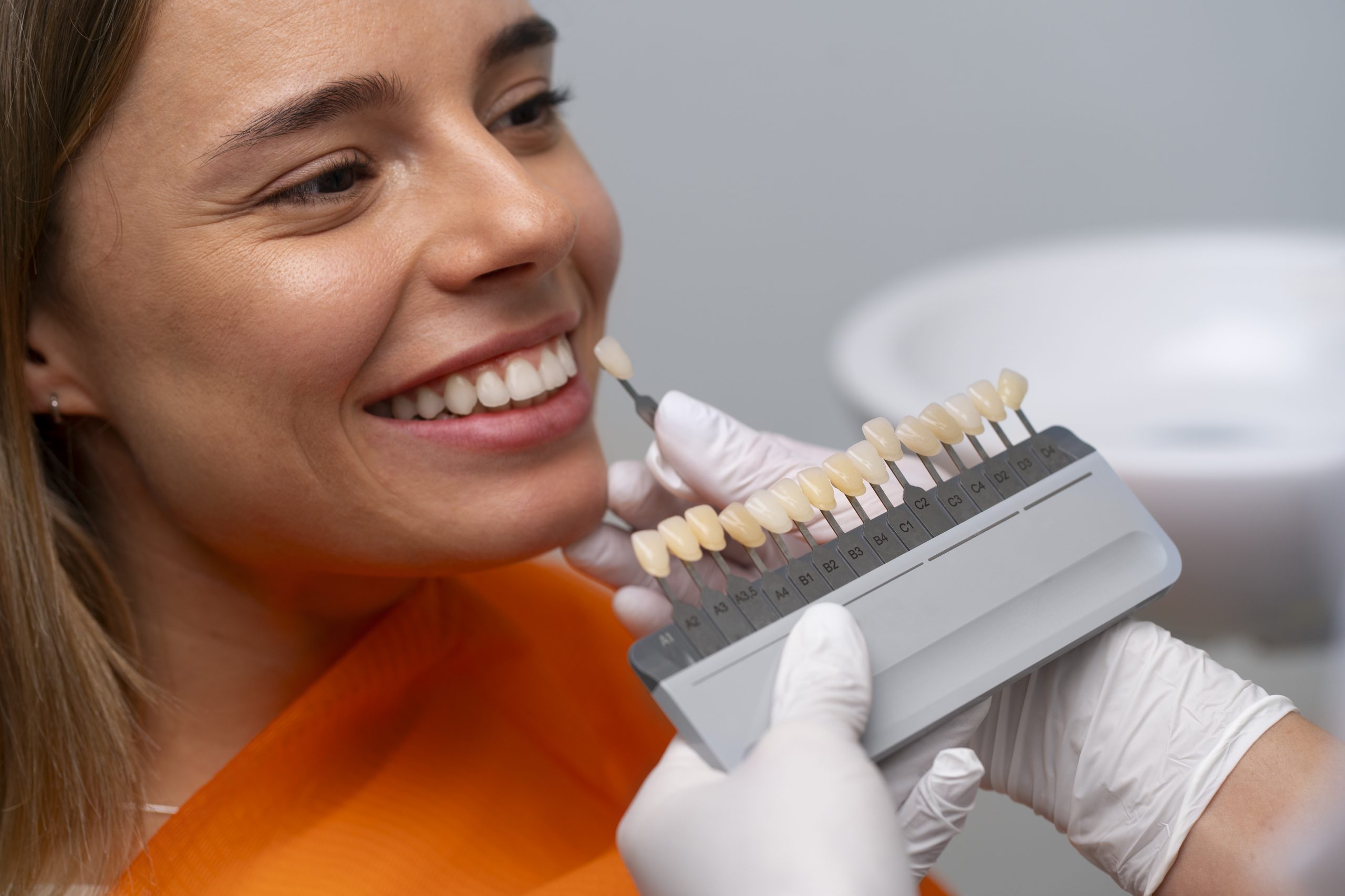 A smiling woman is at a dental clinic, wearing an orange bib. A gloved hand holds a tooth shade guide next to her teeth, suggesting a dental procedure.